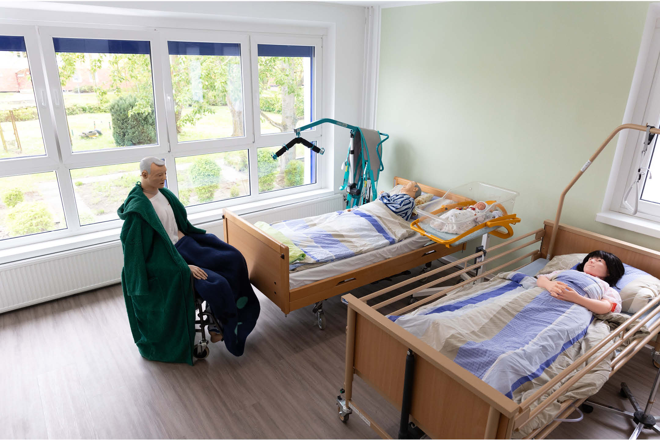 Der Pflegeraum in der bunta, auf dem drei lebensechte Puppen zu sehen sind. Zwei davon liegen zugedeckt in Pflegebetten und einer sitzt im Rollstuhl.
