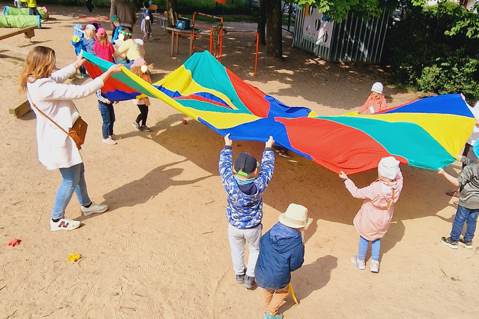 Eine junge Frau in ihren Zwanzigern spielt mit mehreren Kindern mit einem buntem Schwingtuch. Sie befinden sich auf dem Hof eines Kindergartens. Im Hintergrund sieht man weitere Kinder glücklich spielen.